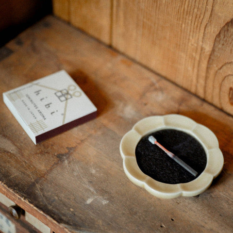 Zbliżenie na drewniany blat, na którym znajduje się opakowanie zapałek zapachowych hibi oraz ceramiczna podstawka z tlącą się zapałką. Zdjęcie oddaje atmosferę prostoty i spokoju, łącząc japońską estetykę z naturalnym, ciepłym tłem. Minimalistyczna kompozycja zachęca do chwili relaksu i uważności.